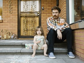 Father sitting on porch with daughter (2-3) and son (6-11 months)