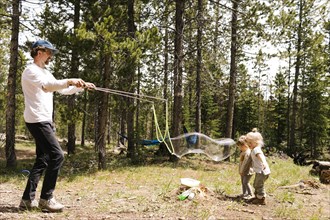 Father making large soap bubbles in forest for two daughters