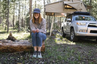 Portrait of smiling woman sitting on log on camping, car with tent in background, Wasatch Cache National Forest