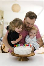 Father carrying kids (2-3, 6-11 months) and blowing birthday candles on cake