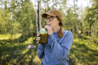 USA, Utah, Uinta National Park, Woman wearing eyeglasses and baseball cap drinking coffee from jar in forest