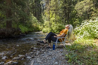 USA, Idaho, Sun Valley, Woman sitting on chair on riverbank in forest