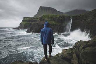 Denmark, Faroe Islands, Gasadalur village, Mulafossur Waterfall, Man standing on cliff and looking at Mulafossur Waterfall falling into ocean