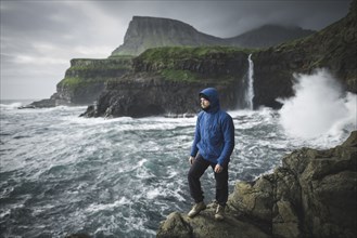 Denmark, Faroe Islands, Gasadalur village, Mulafossur Waterfall, Man standing on cliff with Mulafossur Waterfall in background