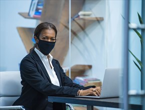 Portrait of businesswoman wearing face mask working on laptop at desk in office