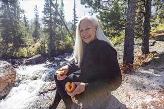 Senior female hiker smiling and cutting fresh peach next to stream