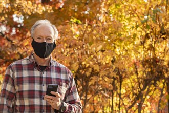 Senior man wearing Covid protective mask and holding smartphone outdoors in fall