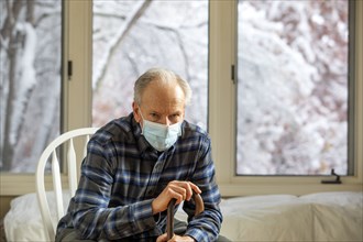 Senior man in Covid protective mask sitting on chair by window and holding cane