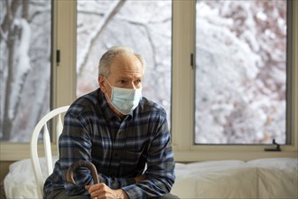 Senior man in Covid protective mask sitting on chair by window and holding cane