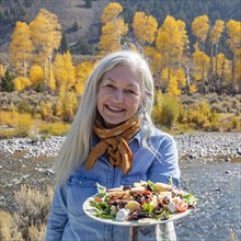 Outdoor portrait of senior woman holding plate of salad while standing along river in autumn