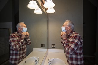 Senior man adjusting protective mask looking in bathroom mirror