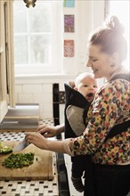 Mid adult mother preparing food with baby son in sling