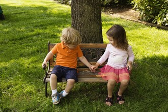 Male and female toddler friends holding hands on scaled park bench