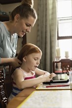 Girl using digital tablet at dining room table whilst mother styling her hair