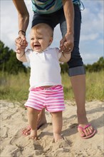 Mid adult woman holding baby daughters hands while toddling in sand