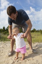 Mid adult man holding baby daughters hands while toddling in sand