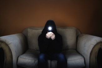 Man in hood sitting on sofa and holding flashlight in front of his face