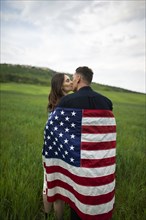 Young couple wrapped in American flag kissing in wheat field
