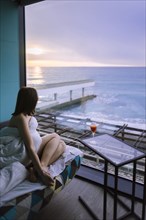 Woman sitting on bed with cocktail in hotel and observing sunrise over sea
