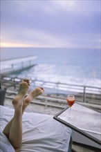 Woman's feet and cocktail in hotel room with view on sea at sunrise