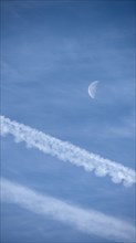 Airplane contrails and waning moon against blue sky