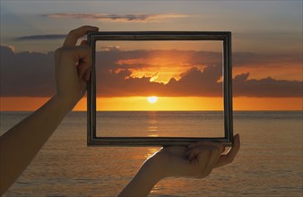Caucasian woman holding frame over dramatic sunset sky and ocean