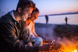 Caucasian couple using cell phone together near fire on beach