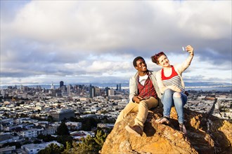 Couple taking cell phone selfie on rock overlooking cityscape