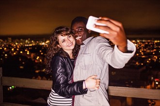 Couple taking cell phone selfie near scenic view of cityscape at night