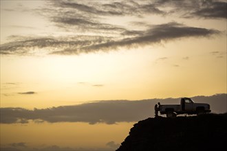 Silhouette of people sitting on pickup truck on cliff at sunset