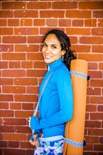 Hispanic woman carrying yoga mat at brick wall