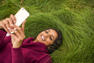 Smiling Indian woman laying in grass texting on cell phone