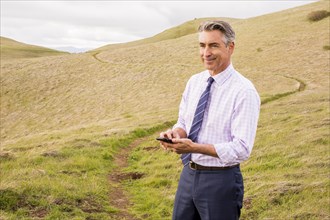 Smiling Caucasian businessman standing in grass holding cell phone