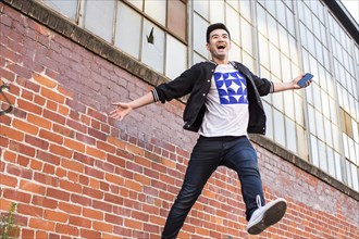 Chinese man jumping for joy near brick wall holding cell phone