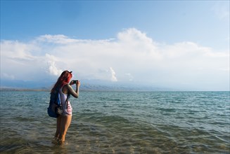 Caucasian woman wading in ocean photographing with cell phone
