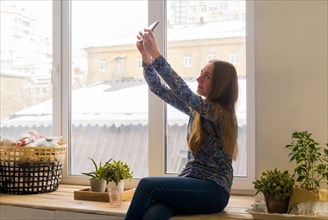 Caucasian woman sitting at window posing for cell phone selfie
