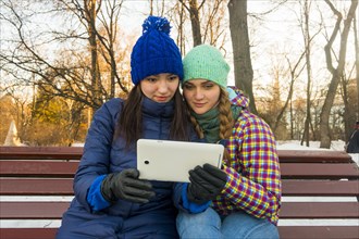 Caucasian women sitting on park bench in the winter holding digital tablet