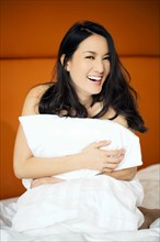 Laughing naked Asian woman kneeling in bed holding pillow