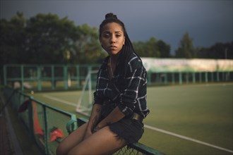Serious African American woman sitting on fence at sports field
