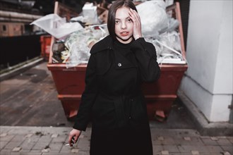 Caucasian woman wearing black coat smoking cigarette near dumpster