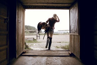 Caucasian woman carrying horse saddle in barn doorway