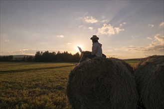 Mari man sitting on hay bale at sunset using laptop