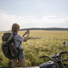 Caucasian woman standing in field near motorcycle using cell phone
