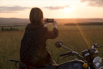 Caucasian woman with motorcycle in field photographing sunset