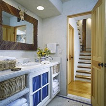 View of rustic wood stairway through open door on right. White open cabinets on left with wicker baskets holding white cotton greek  key pattern towels. White ceramic tile and yellow calla lilies. Rus...