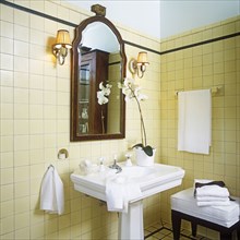 1920s style. Detail of white stately pedestal sink in center and mahogany framed mirror. White orchids sitting on right side of sink. Shade sconces. Cream