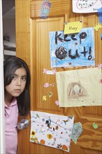Hispanic girl peering out bedroom door with Keep Out signs