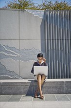 Asian businesswoman sitting on concrete wall using laptop