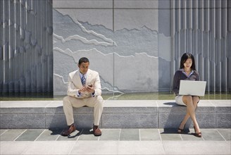 Business people sitting on concrete wall outdoors using technology