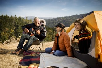 Women at camping tent listening to man playing guitar